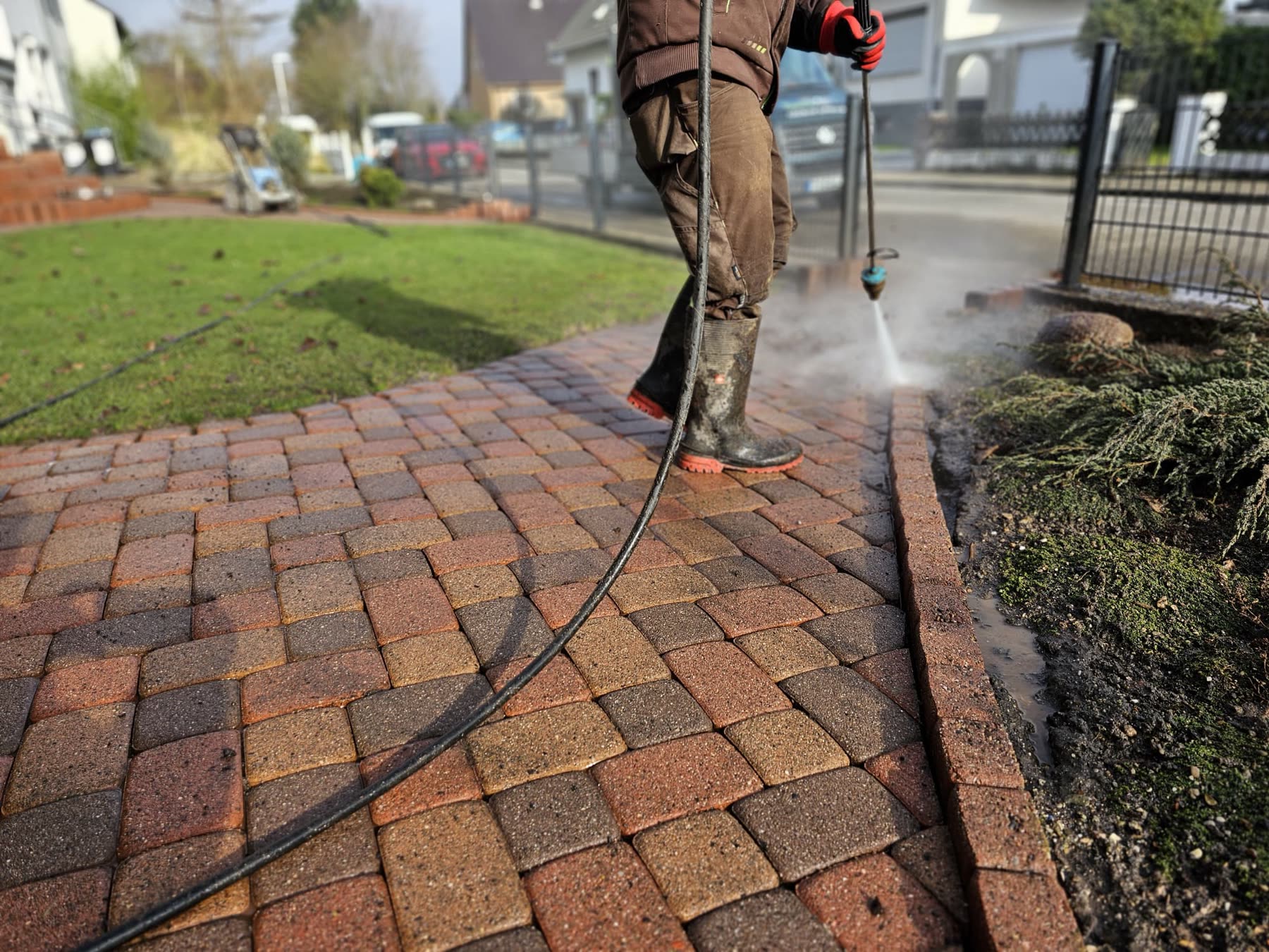 Reinigungskraft führt einen Hochdruckstrahler über einen Pflasterweg, Wasser spritzt auf